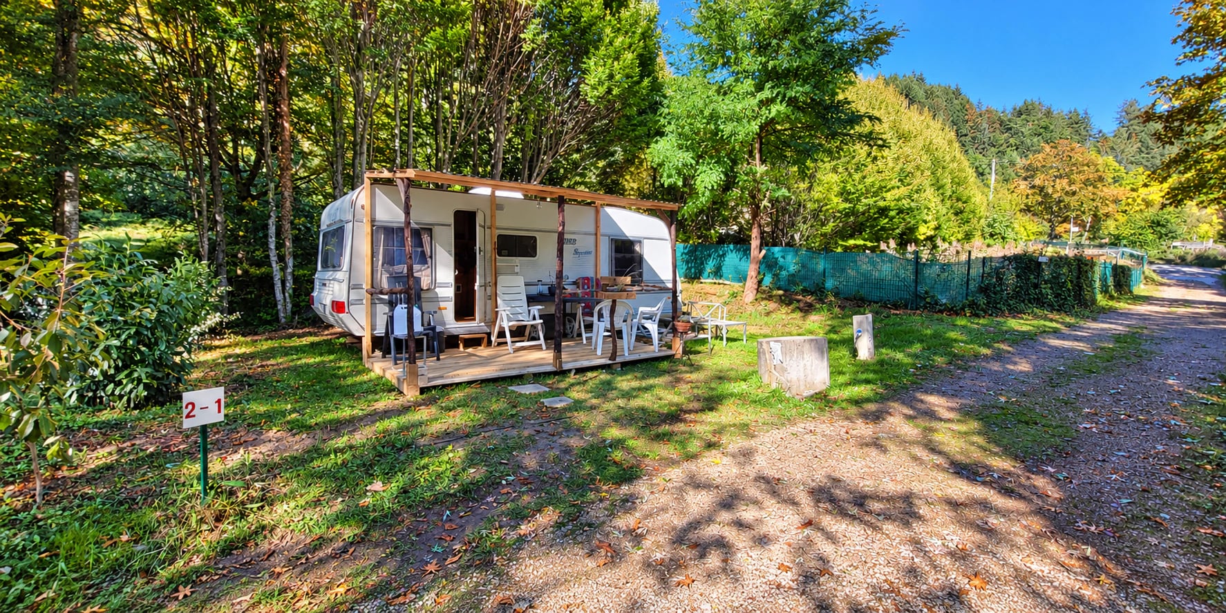 De vaste caravan met overdekt houten terras, op een plek tussen de bomen